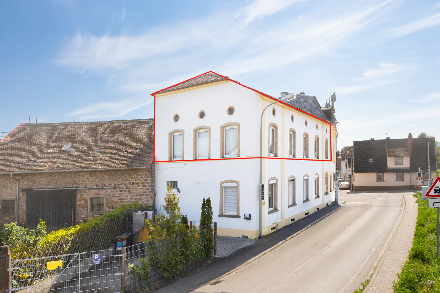 Two-story white building with a red roof outline, next to a stone building and a road.