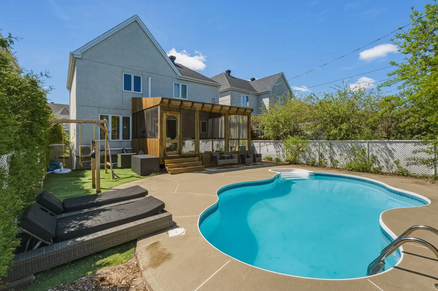 Backyard view of a two-story house with a blue pool, lounge chairs, and a wooden patio under a sunny sky.