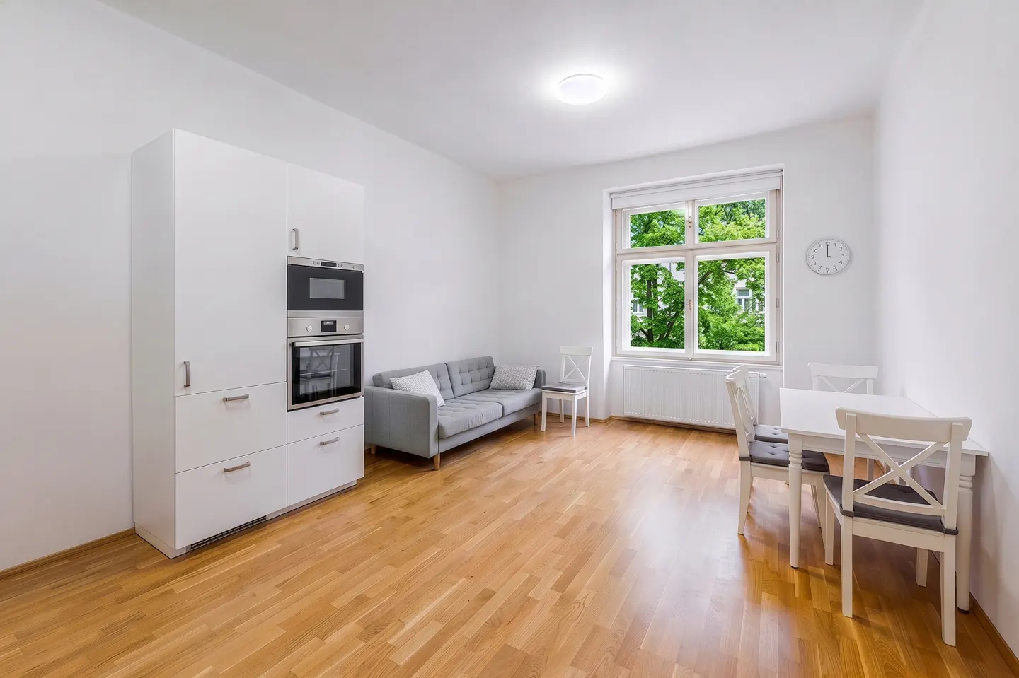 Bright, modern apartment interior with wood floors, white walls, a gray sofa, and a white dining set near a window with green trees outside.