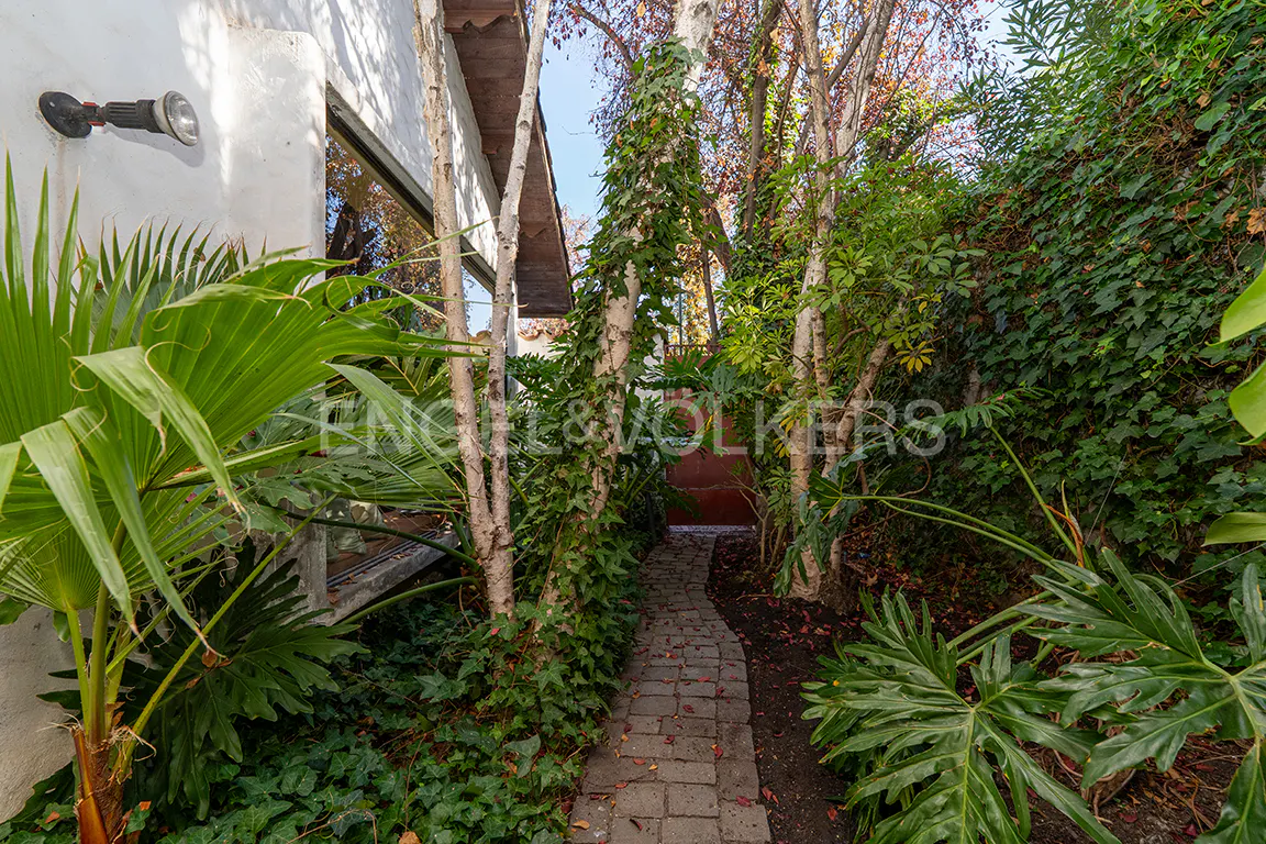 A brick path winds through a lush garden with trees, ivy, and tropical plants. A white building with a light fixture is visible on the left.