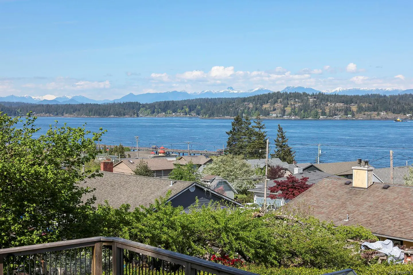 Scenic view from a deck overlooking rooftops, a blue bay, forested islands, and snow-capped mountains under a bright blue sky.
