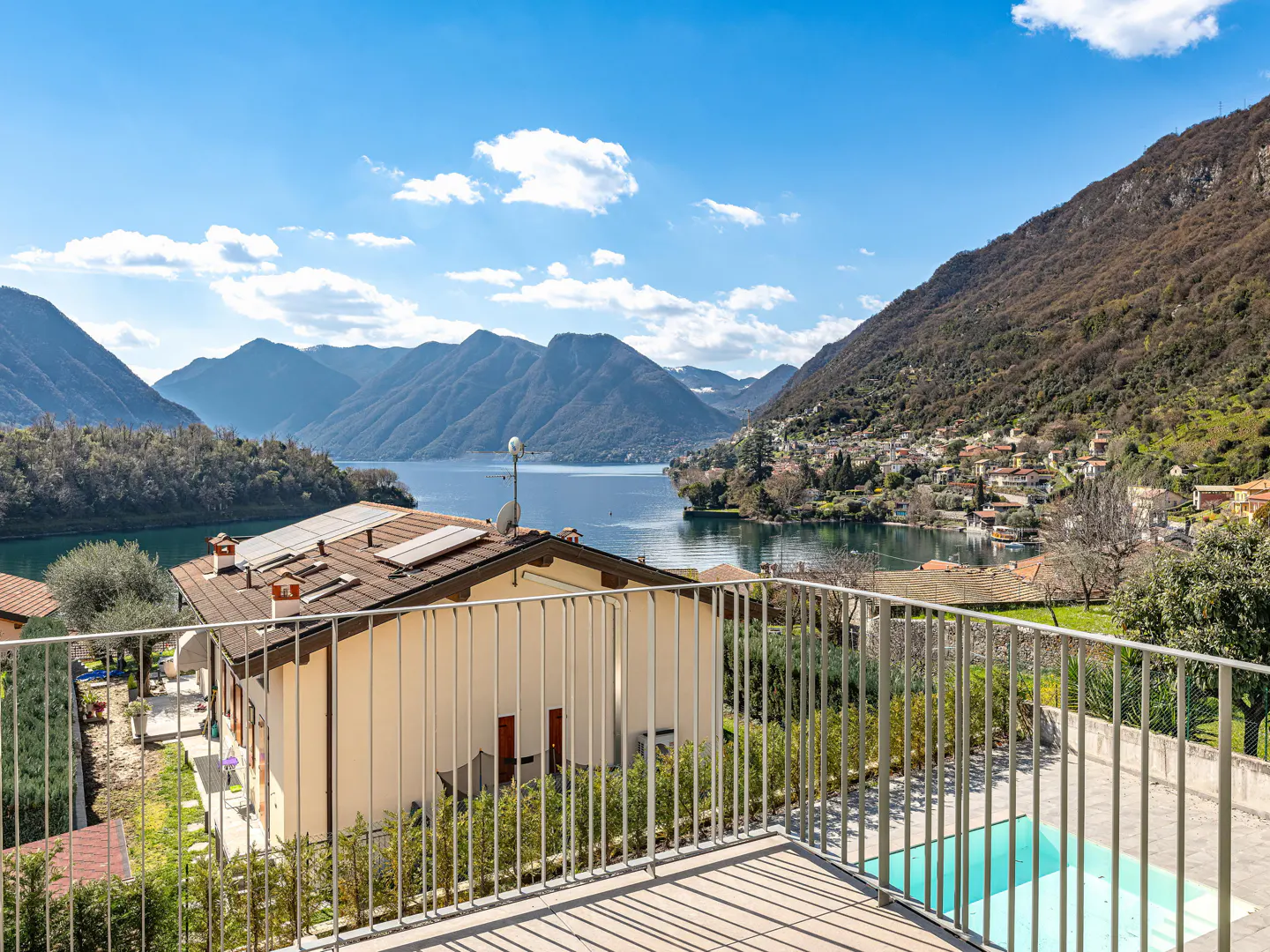 View from a balcony overlooking a lake, mountains, and a house with a pool on a sunny day.