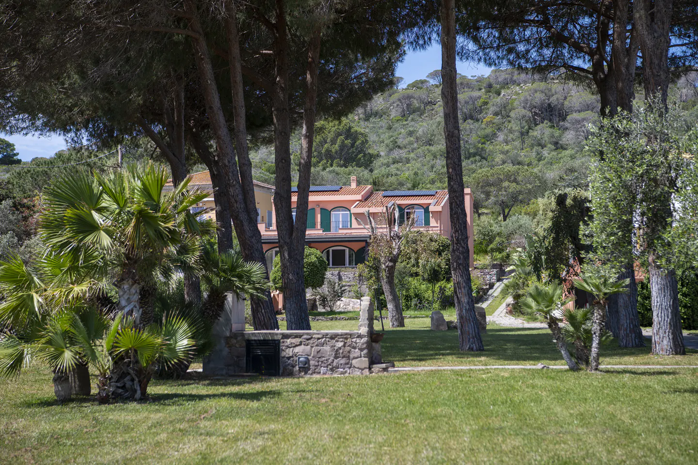 A salmon-colored house with green shutters and a red tile roof, partially obscured by trees, sits on a green lawn.