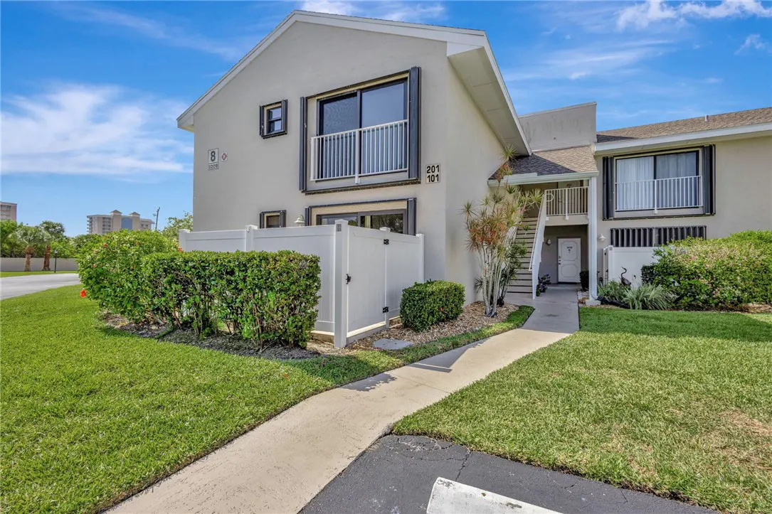 Two-story condo with a white fence, green bushes, and a sidewalk leading to the entrance. Balcony on the second floor. Blue sky.