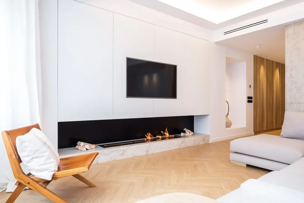 Modern living room with a white wall unit, TV, linear fireplace, and a tan leather chair with a white pillow.
