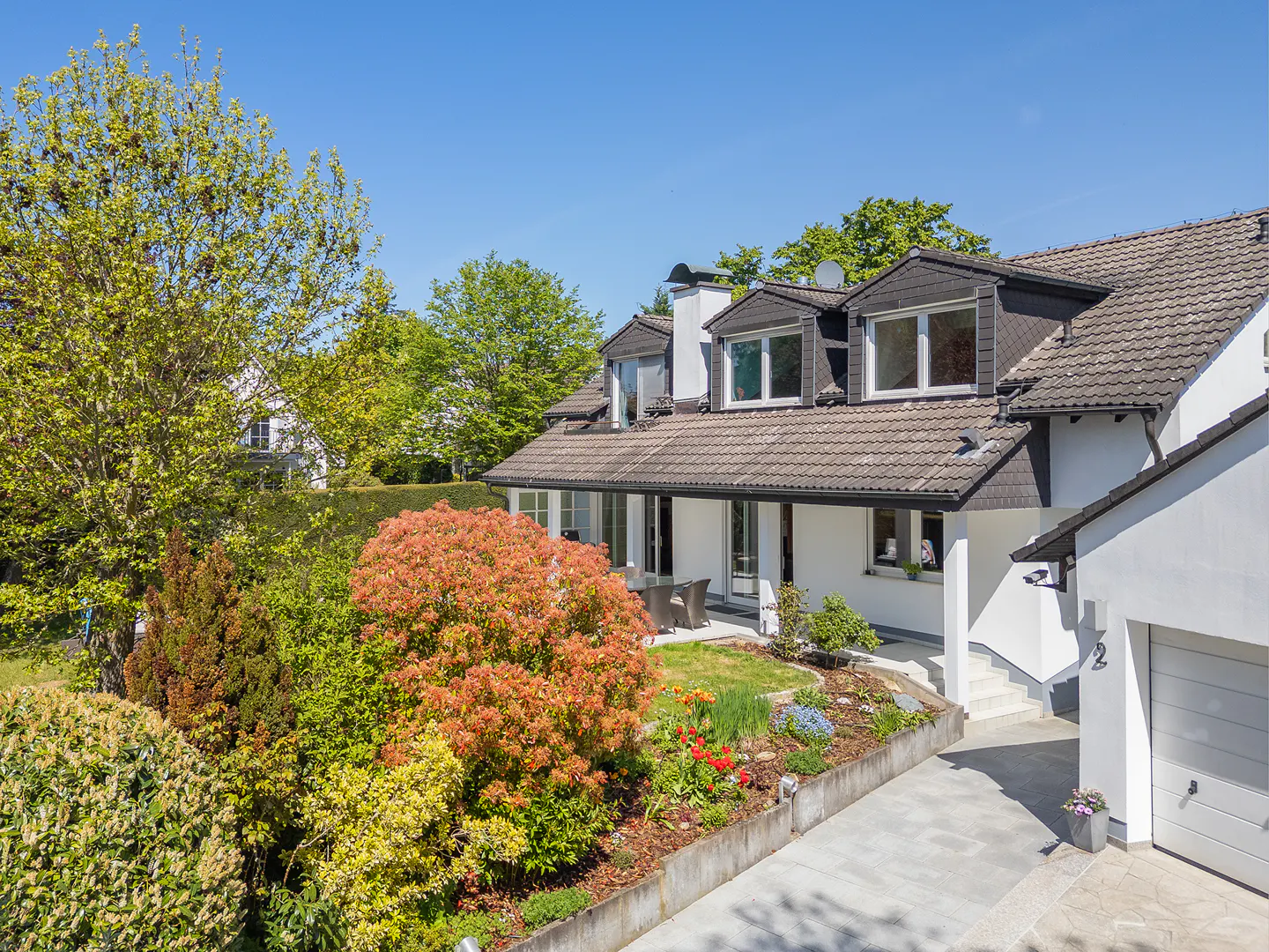 Exterior view of a two-story white house with a gray roof, dormer windows, and a lush garden with colorful flowers.