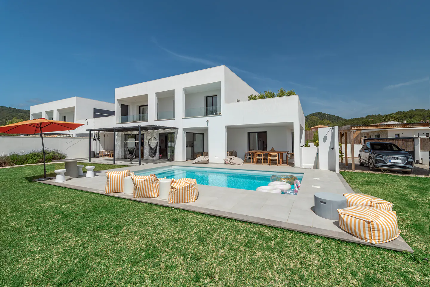 Modern white house with a pool, striped cushions, and orange umbrella on green lawn under a blue sky.
