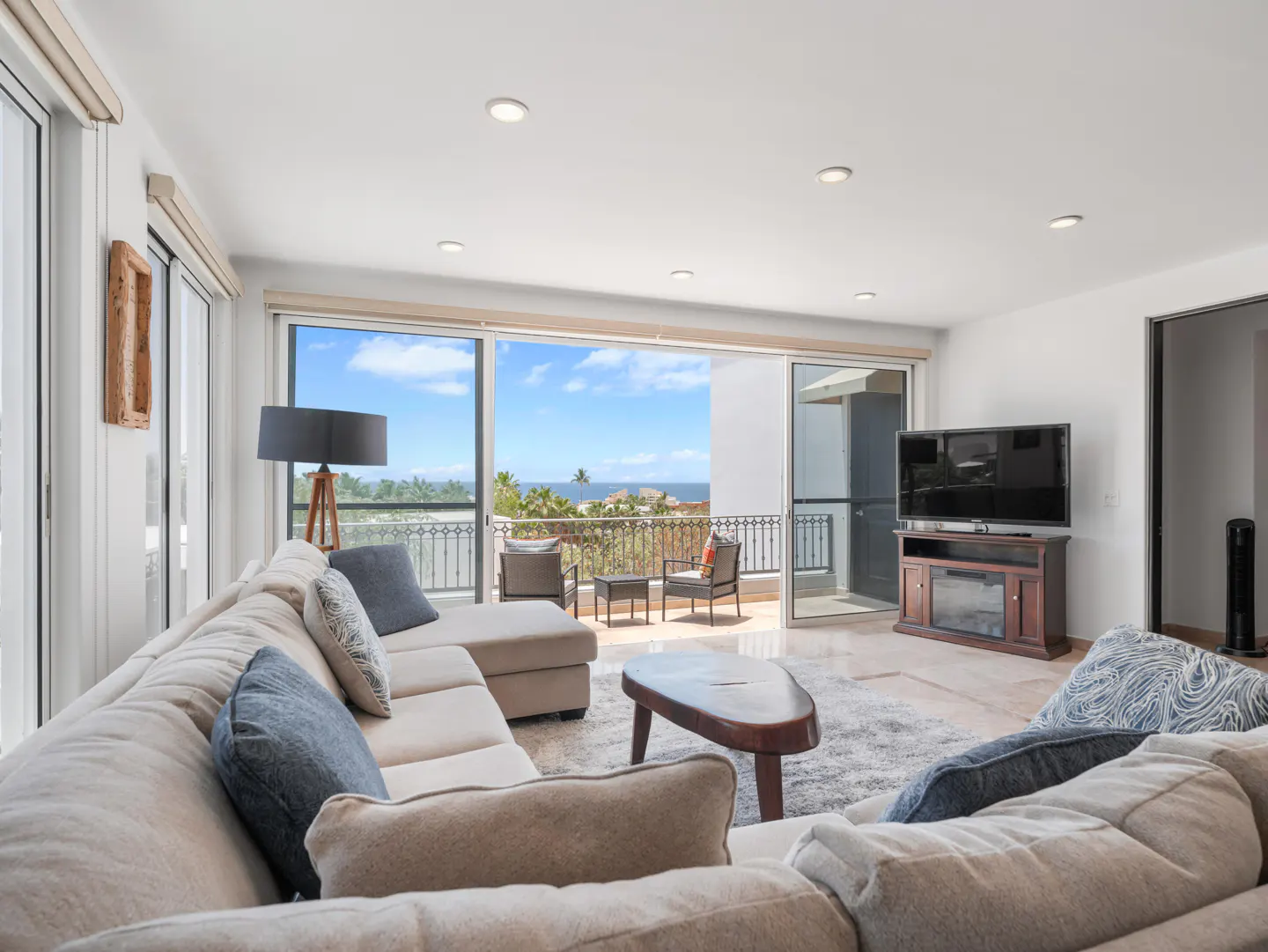 Bright living room with a large sectional sofa, coffee table, TV, and sliding doors opening to an ocean-view balcony.