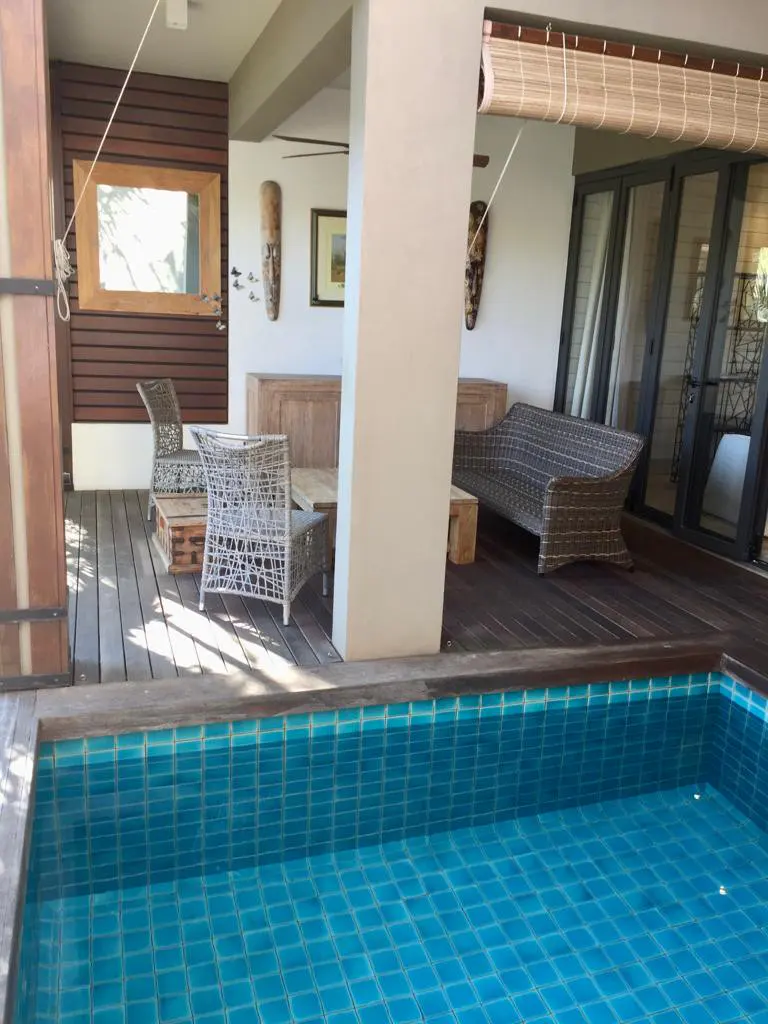 Outdoor patio with a blue-tiled pool, wicker furniture, and wooden accents. The patio has a covered area with a white pillar and decorative masks on the wall.