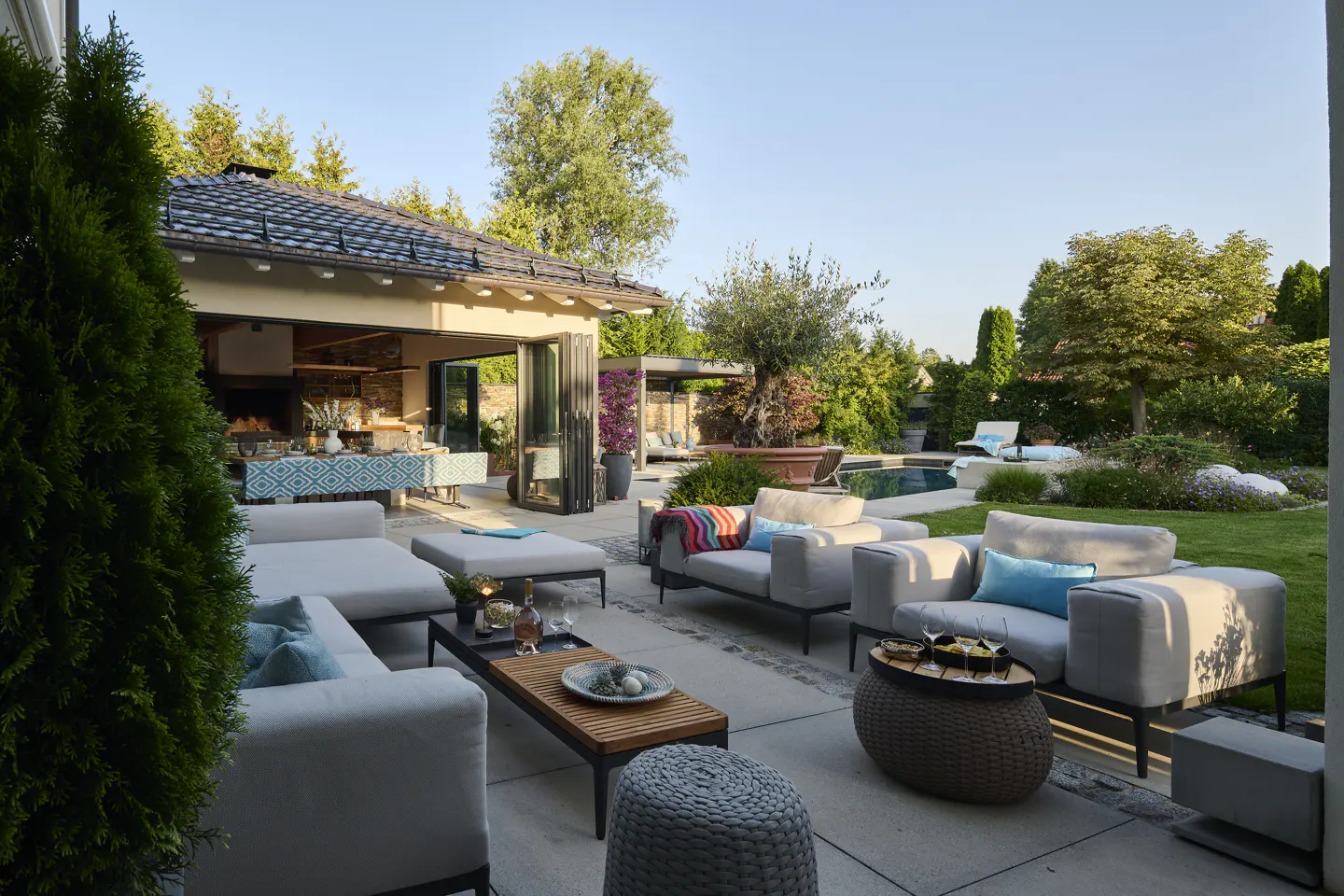 Outdoor patio with gray furniture, blue pillows, and a wooden coffee table. A pool and lush greenery are in the background.