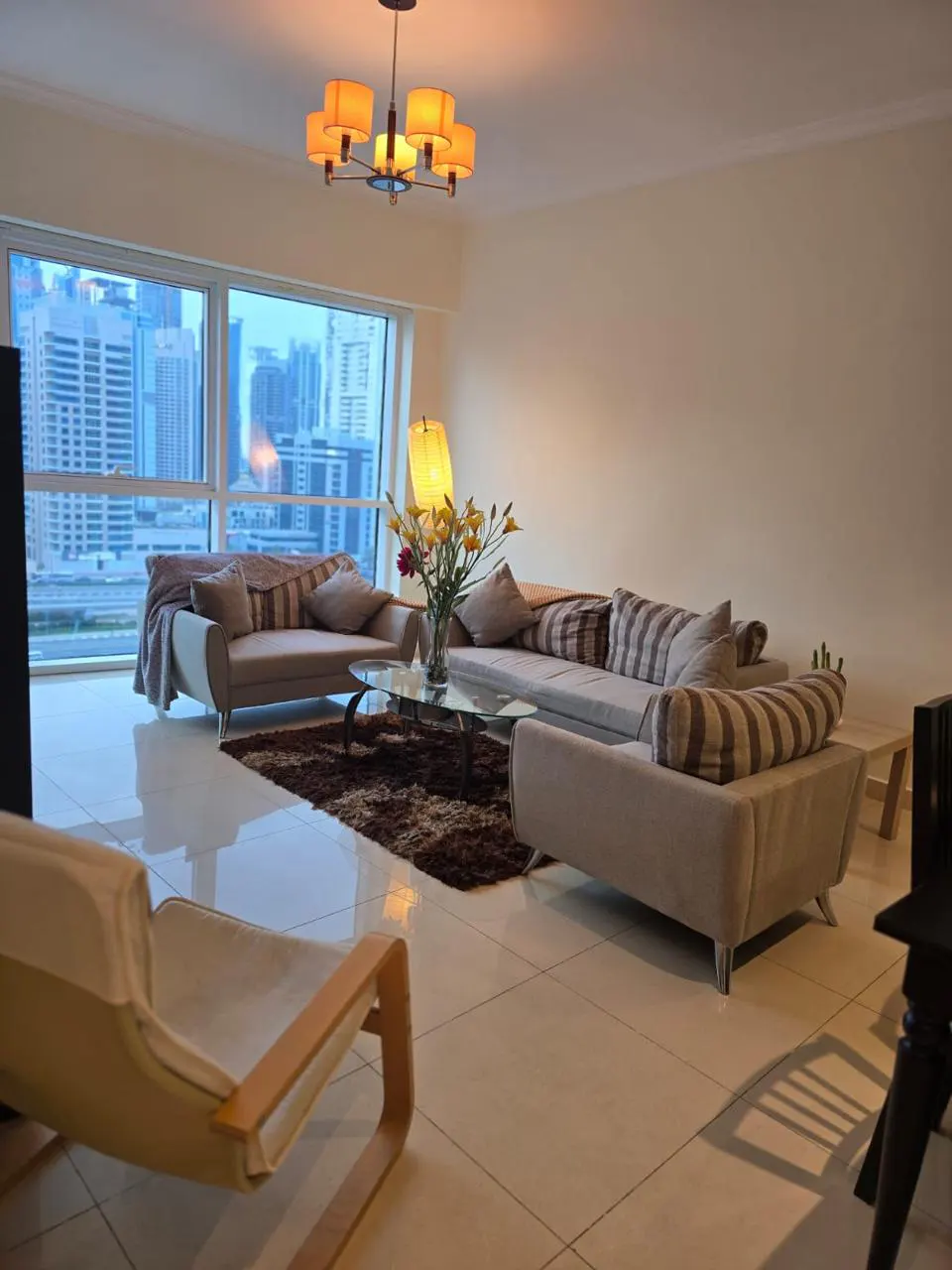 Living room with beige sofas, brown rug, glass table, and city view through large windows. A modern chandelier hangs above.