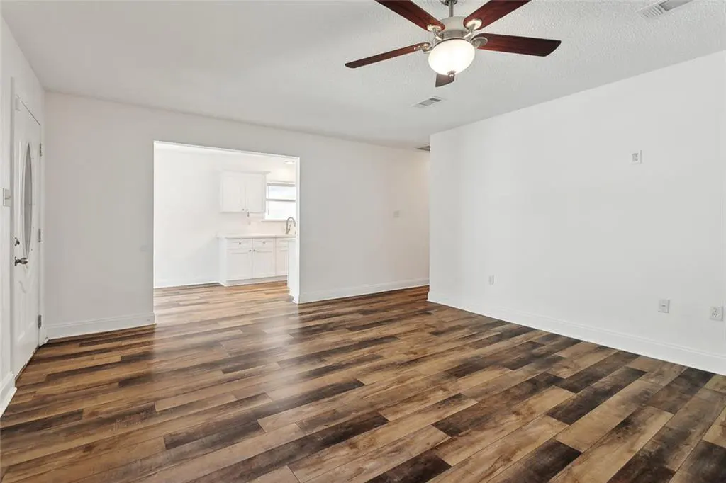Bright, empty living room with wood floors and white walls. A ceiling fan hangs above, and a glimpse of the kitchen is visible through an opening.