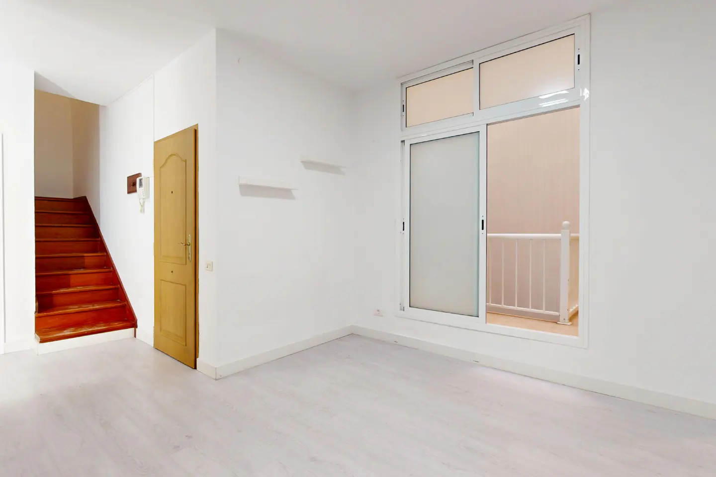 Bright, empty room with white walls, light wood floors, and a sliding glass door. A wooden door and stairs are on the left.