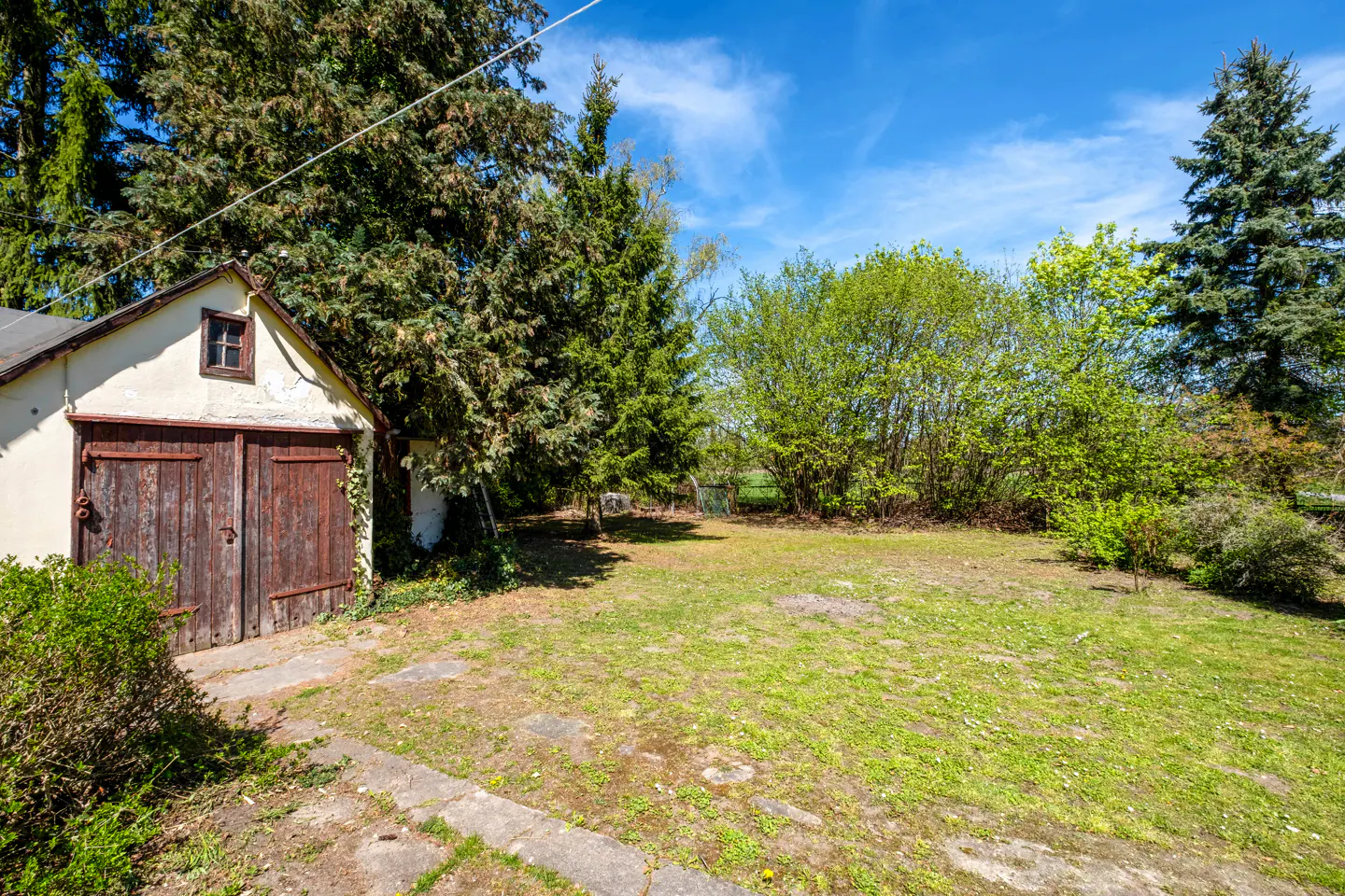 A small, cream-colored garage with dark wood doors sits on a green lawn under a blue sky with trees.