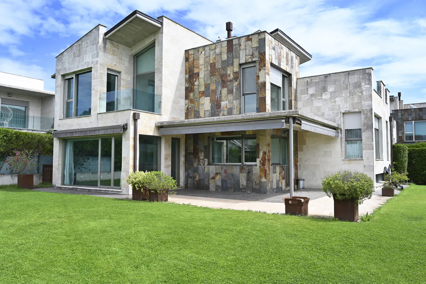 Modern two-story house with stone and stucco exterior, large windows, and green lawn under a blue sky.