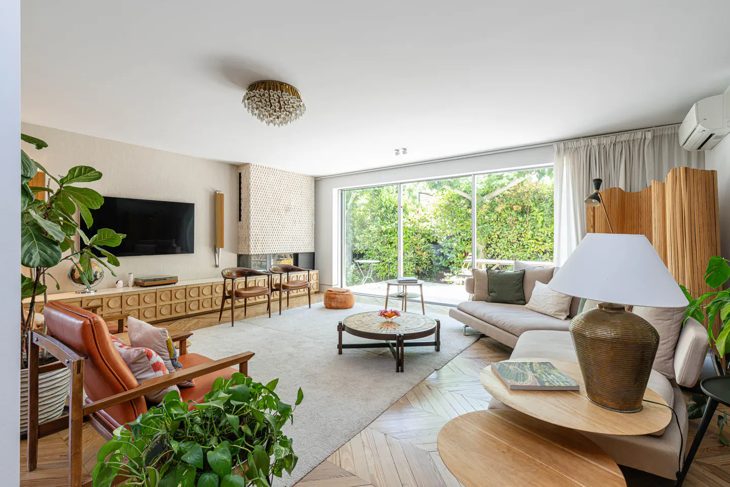 Bright living room with a fireplace, large windows overlooking a green garden, and a mix of modern and vintage furniture.