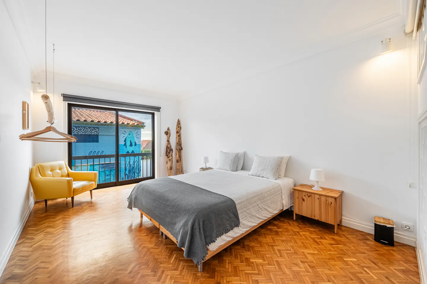 Bright bedroom with herringbone wood floors, white walls, a bed with a gray blanket, and a yellow chair near a sliding glass door.