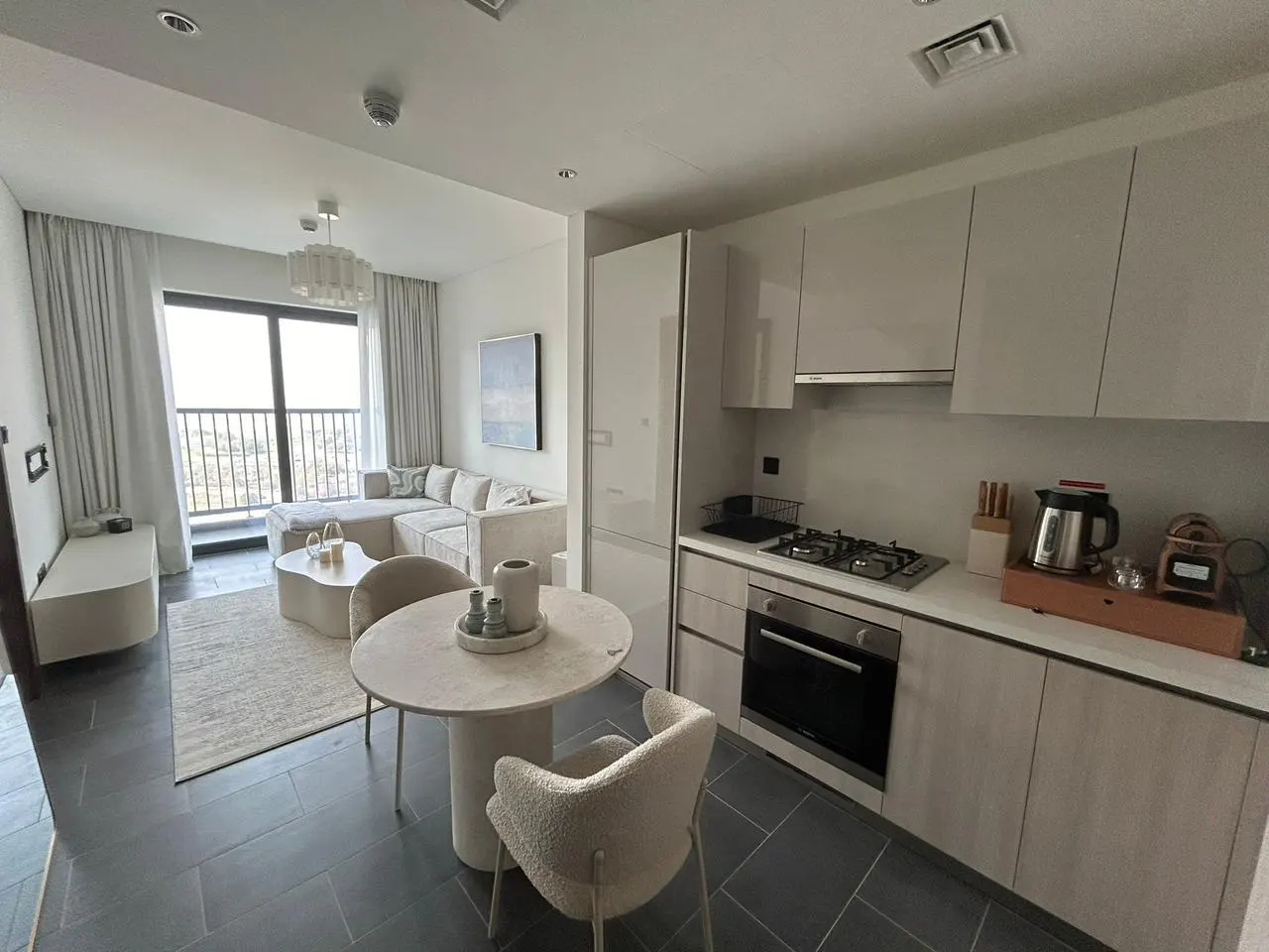 A bright, modern apartment with a living room, dining area, and kitchen. A beige sofa faces a window with a city view. A round table with two chairs sits near the kitchen.