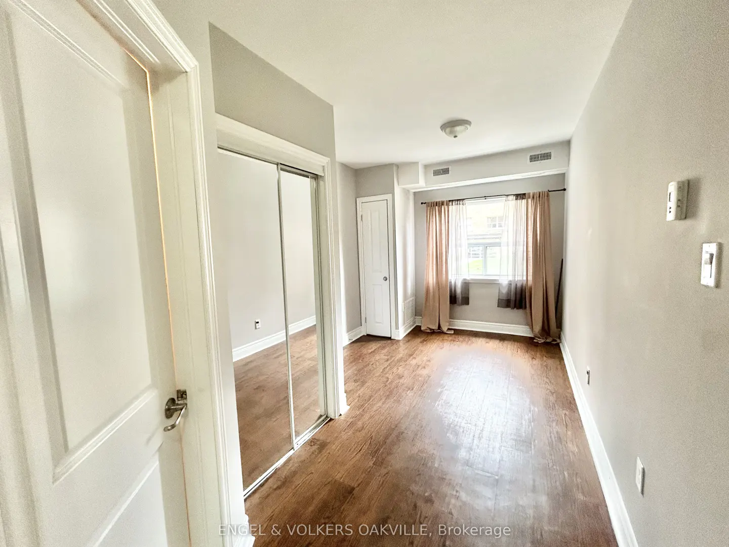A bright, empty room with hardwood floors, light gray walls, and a window with beige curtains. A mirrored closet and white door are visible.