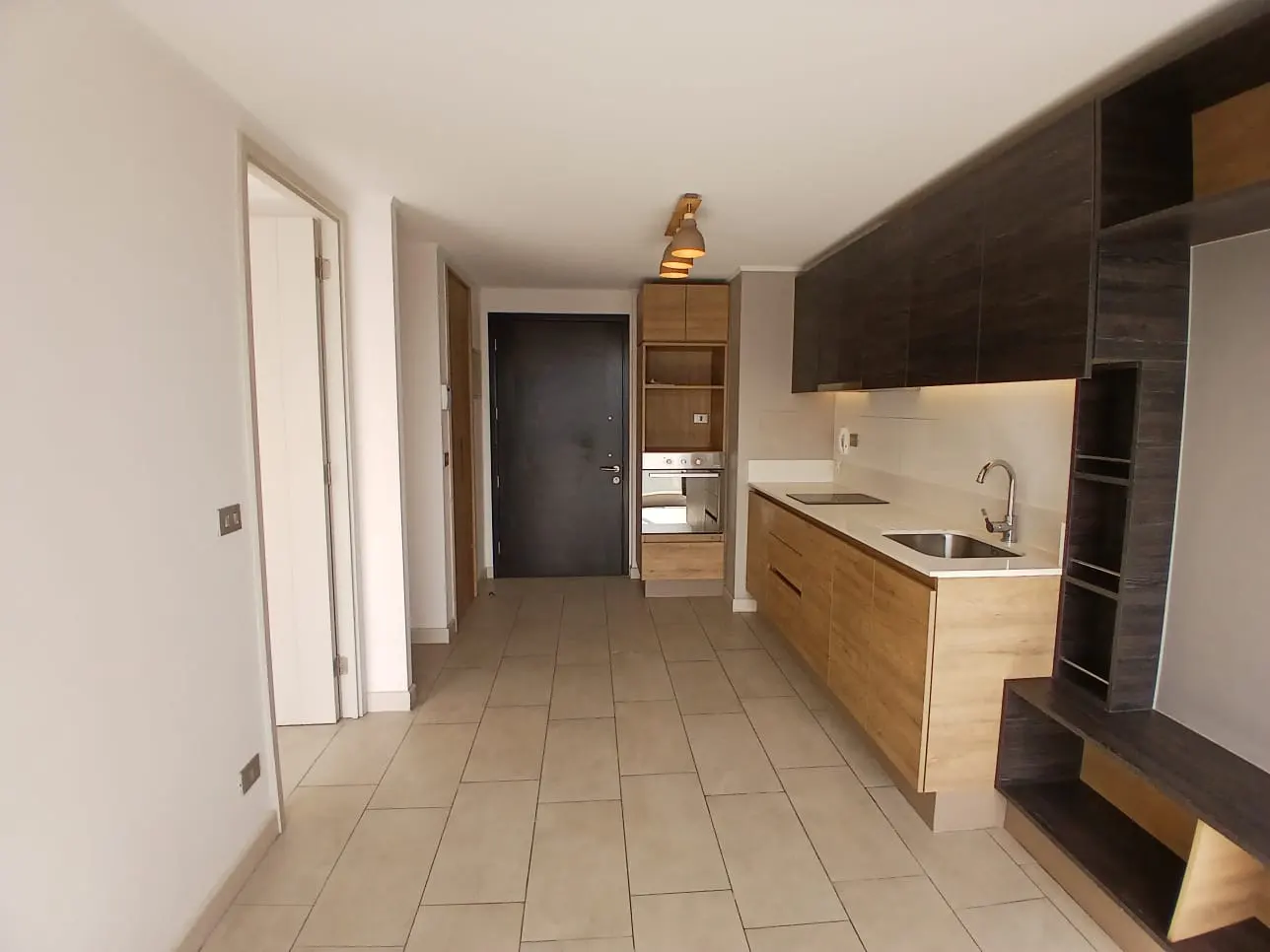 A modern, empty apartment with light tile floors, white walls, and a kitchen with wood cabinets and dark upper cabinets.