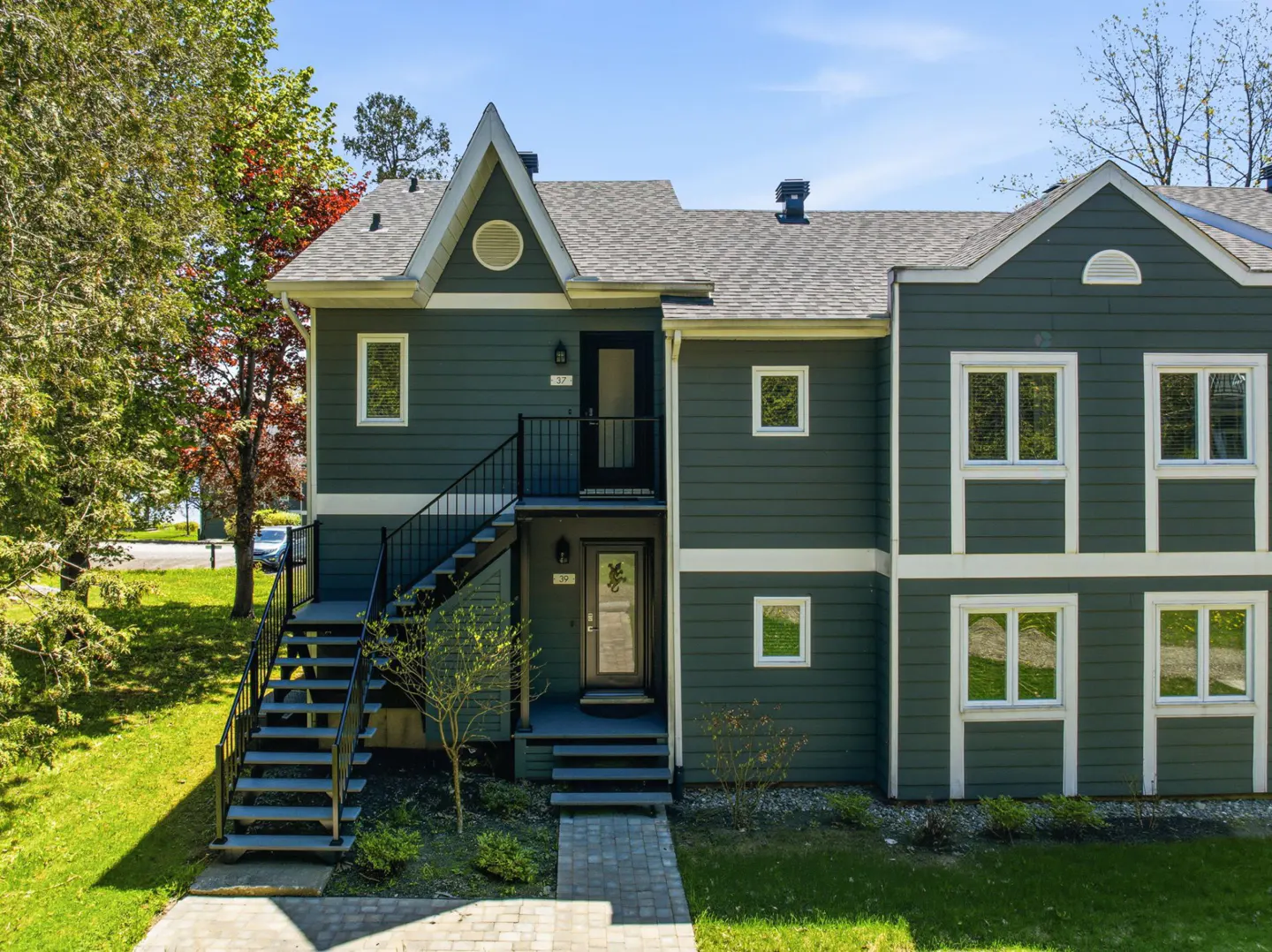 Two-story townhouse with dark green siding, white trim, and a gray roof. Black stairs lead to the upper unit. A brick walkway leads to the front door.