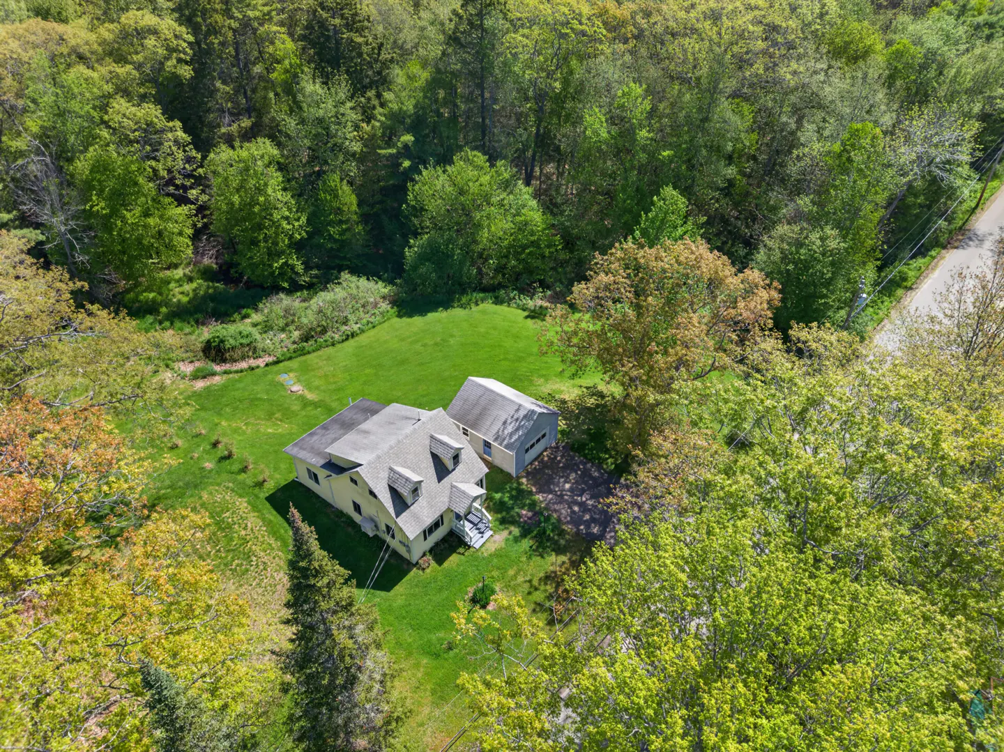 Aerial view of a light-colored house and detached garage surrounded by a lush green lawn and dense forest.