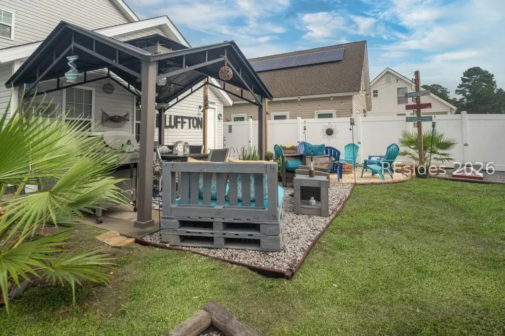 Backyard patio with gray pallet furniture, blue cushions, and a black metal pergola. A white fence and green lawn complete the scene.