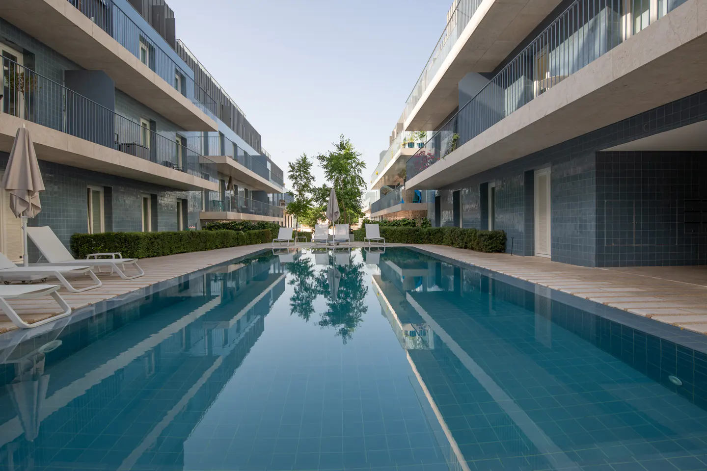 A long, blue swimming pool between two modern apartment buildings with balconies.