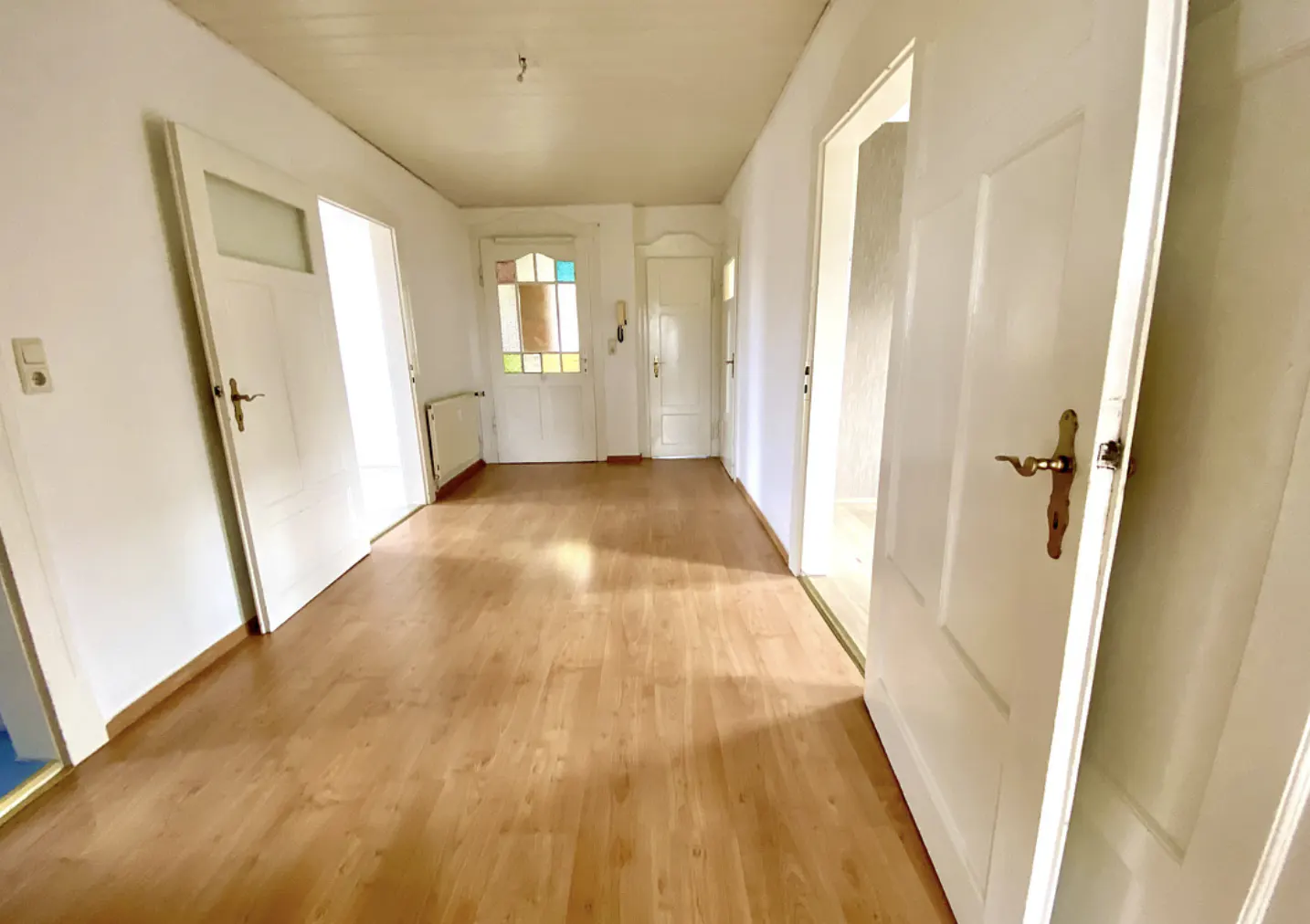 A bright hallway with wood floors and white walls. Several white doors are visible, some open. A radiator is on the left wall.
