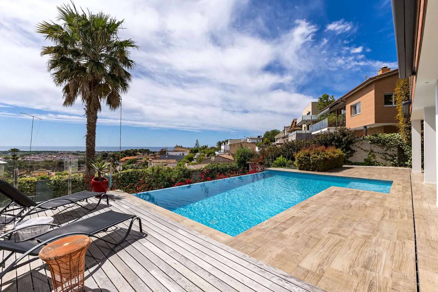 Outdoor pool with blue water, surrounded by a wooden deck, lounge chairs, and a palm tree. City and ocean views in the background.