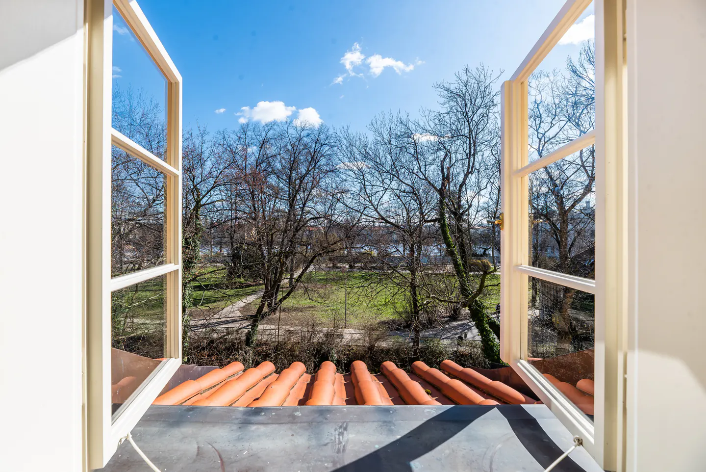View from an open window with a white frame, overlooking a park with bare trees and a blue sky with clouds. Orange roof tiles are visible below.