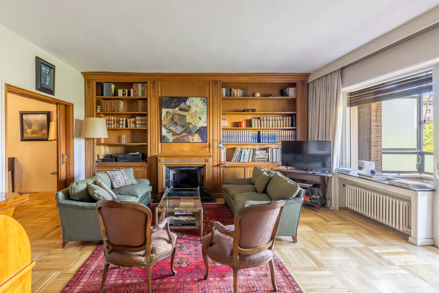 Living room with wood floors, built-in bookshelves, and a red rug. Two green sofas face each other, with two chairs in front. A fireplace is centered.