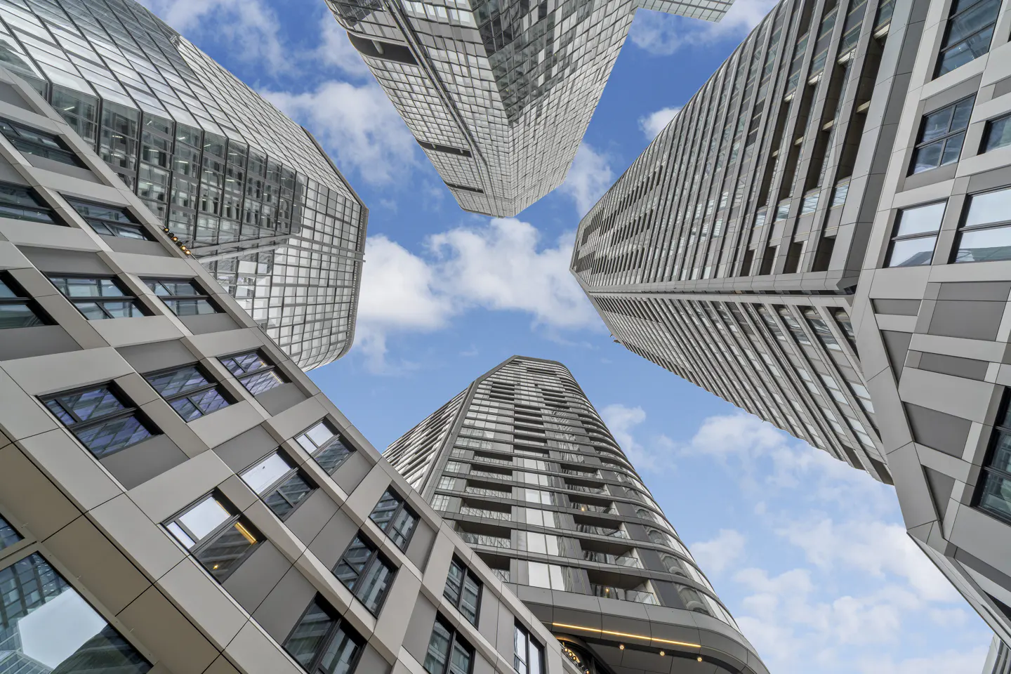 Low-angle view of four modern glass and grey steel skyscrapers reaching toward a blue sky with scattered white clouds.