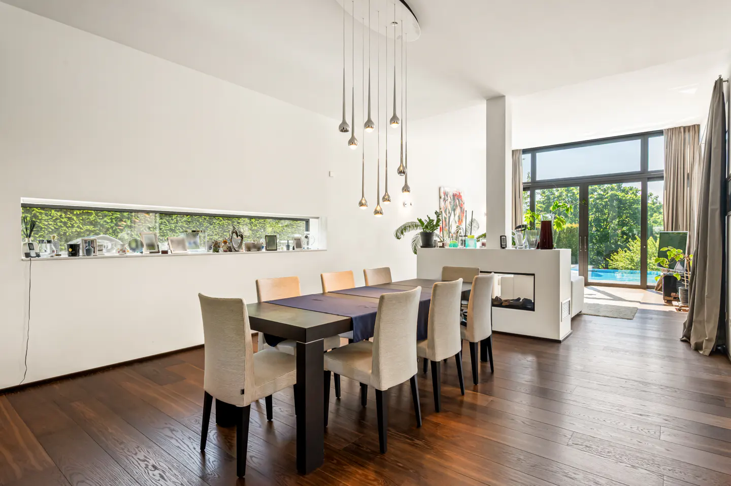 Modern dining room with dark wood floors, a long table with beige chairs, pendant lights, and a view of a backyard pool.