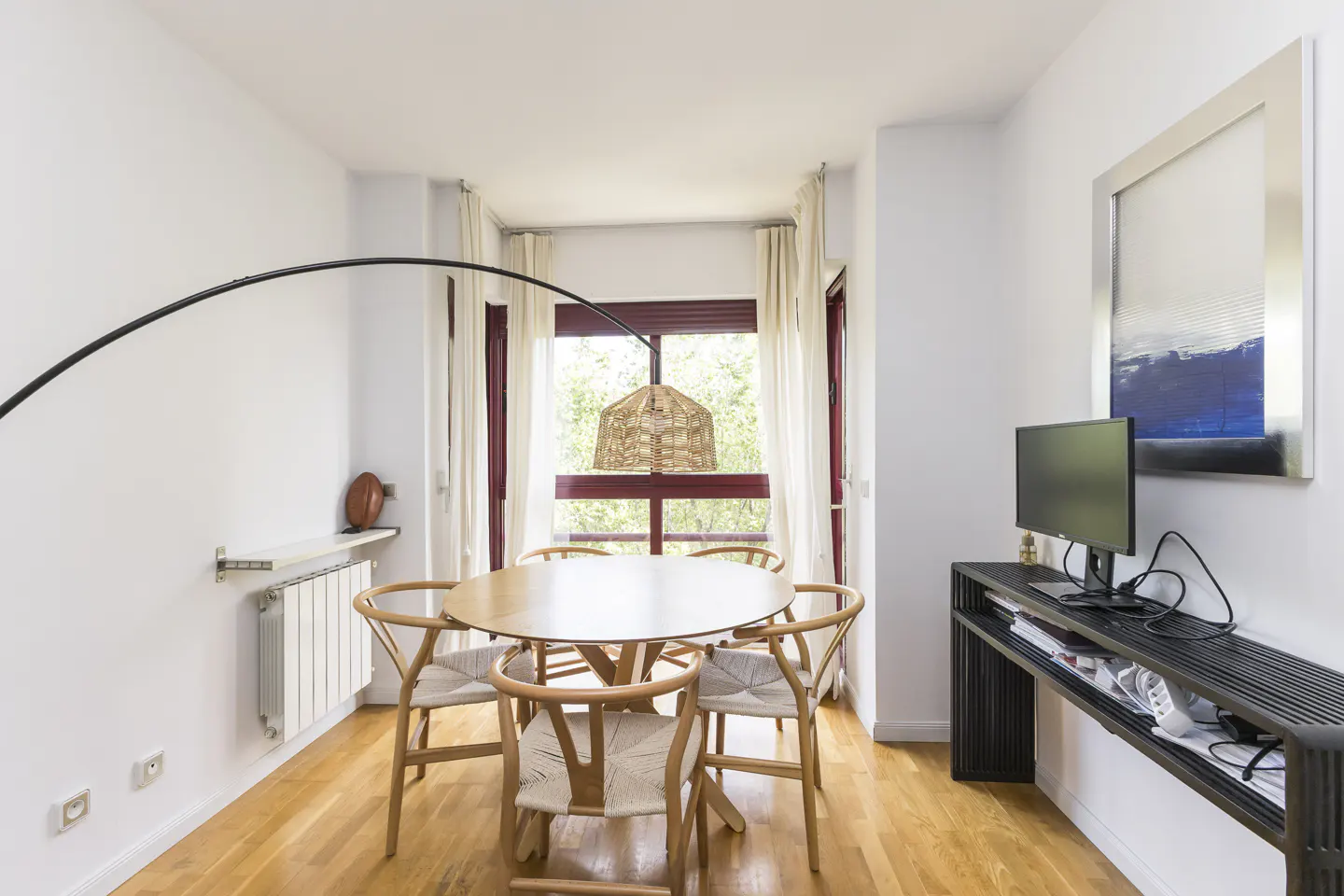 Bright dining room with wood floors, white walls, and a round table with six chairs. A black console table and monitor are against the wall.
