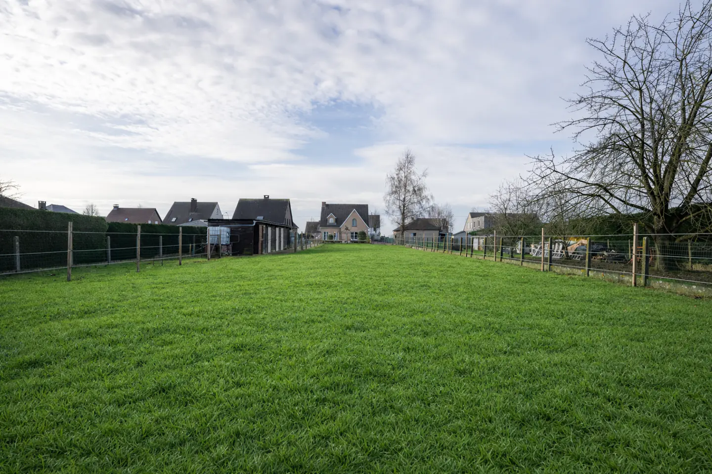 A large, fenced green grass field leading toward a residential neighborhood under a cloudy sky.