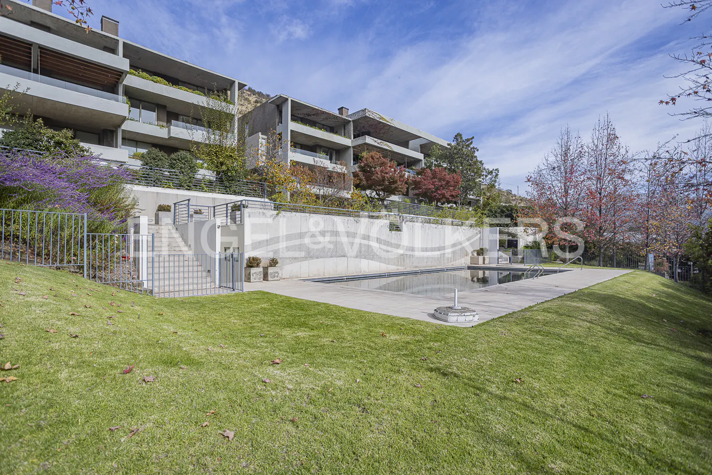 Modern apartment building with balconies overlooking a swimming pool, green lawn, and autumn trees under a blue sky.