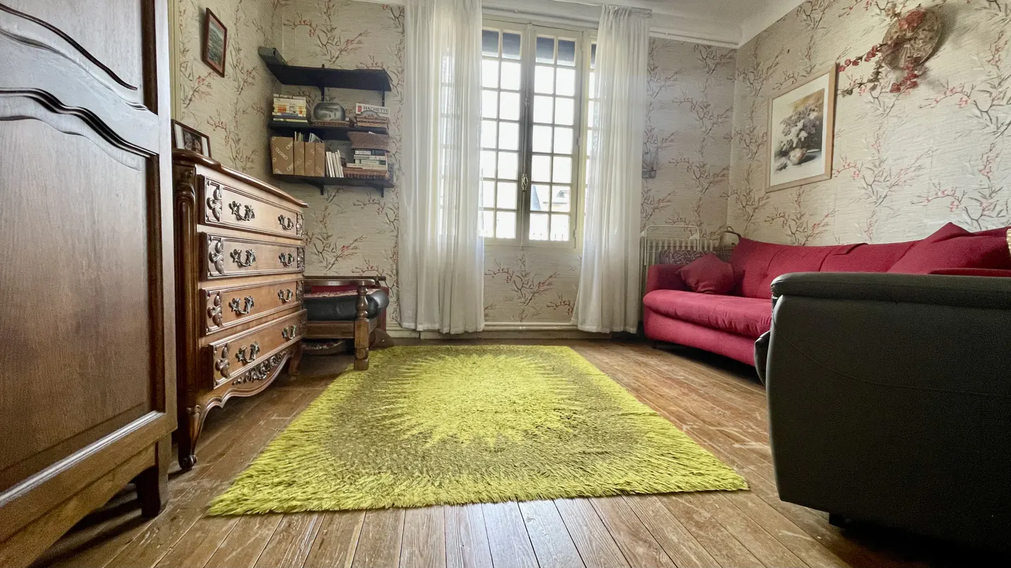 A cozy room with floral wallpaper, a red sofa, and a bright green rug on a wood floor. A dresser and window with white curtains are also visible.