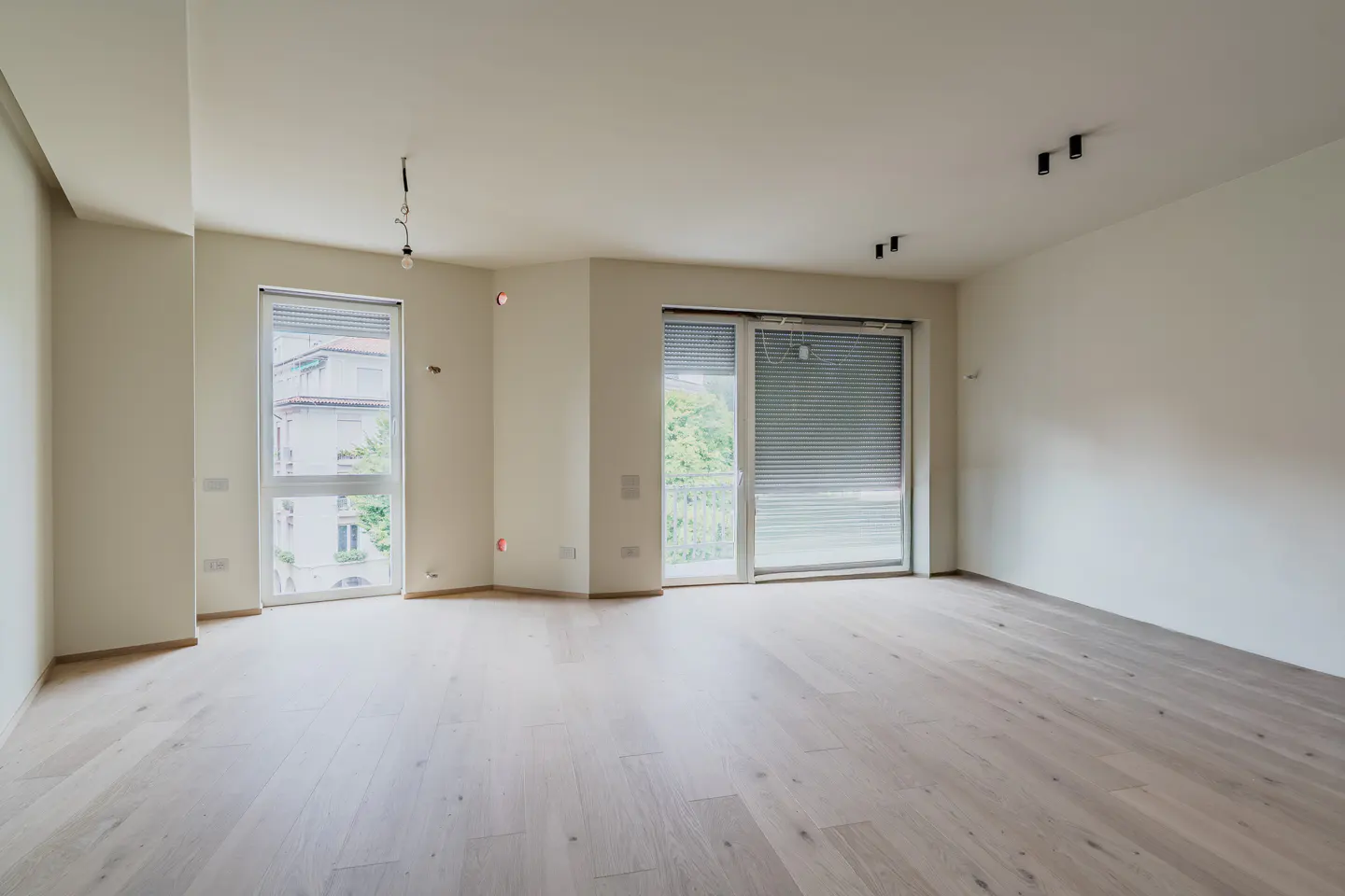 Empty, bright living room with light wood floors, white walls, recessed ceiling lights, and large windows with blinds.