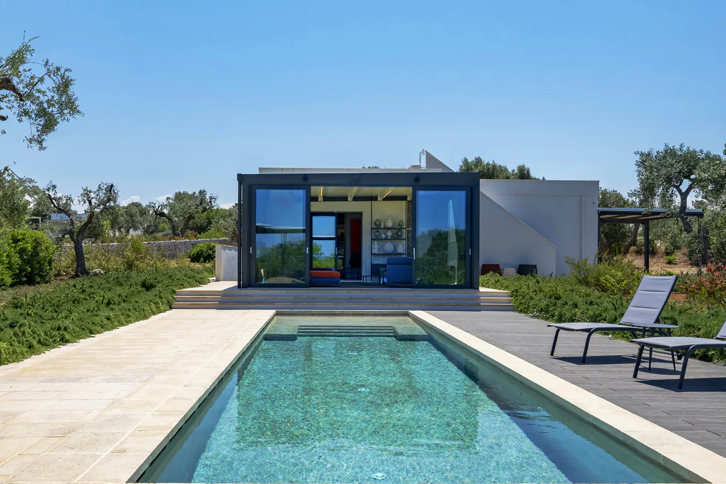 Modern home with glass walls and a rectangular pool, surrounded by lush greenery and lounge chairs under a clear blue sky.