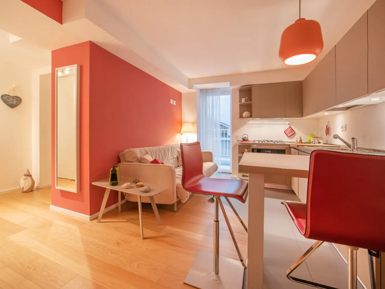 Bright apartment interior with wood floors, red accent wall, and open kitchen. A white sofa and red bar stools add pops of color.