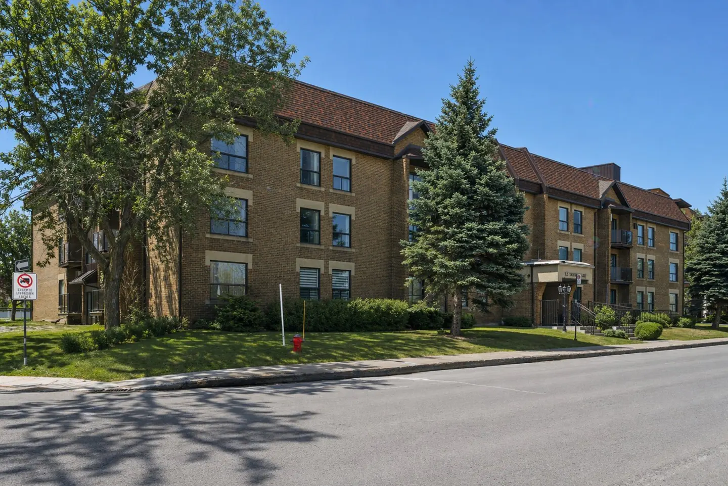 Three-story brown brick apartment building with a dark shingled roof, surrounded by green lawns and evergreen trees.