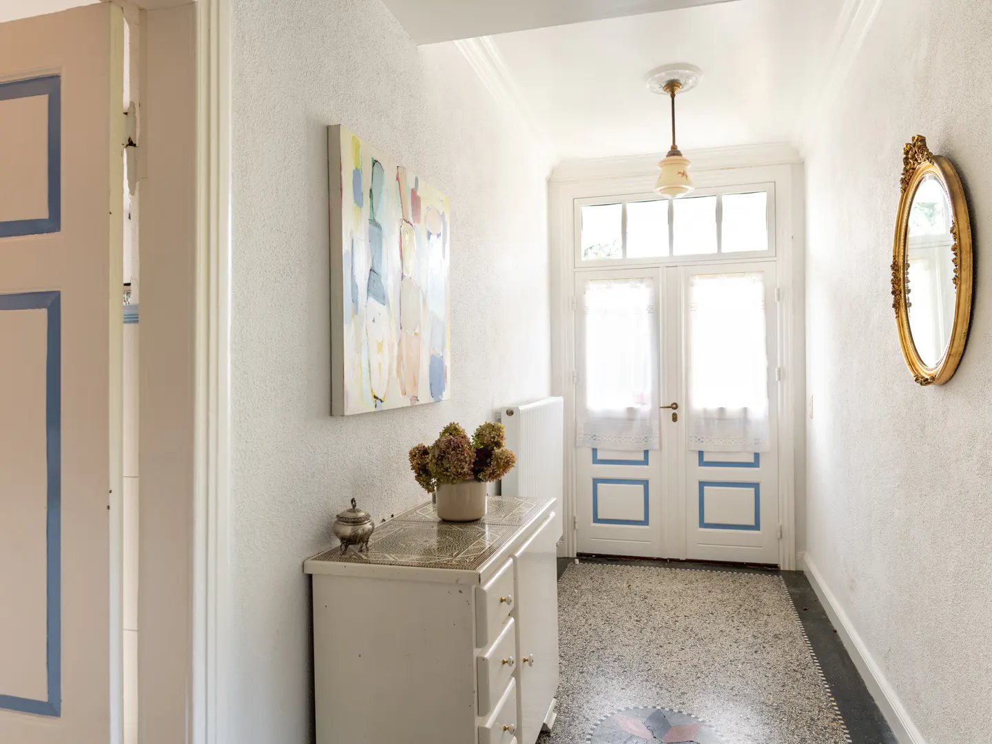 Bright hallway with white walls, a terrazzo floor, and a white cabinet with a vase of flowers. A gold-framed mirror hangs on the wall.