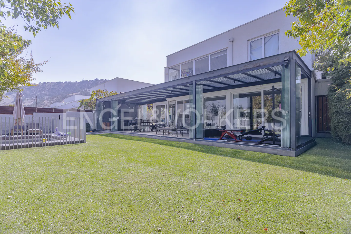 A modern white house with a glass-enclosed patio and green lawn. Exercise equipment is visible inside the patio.