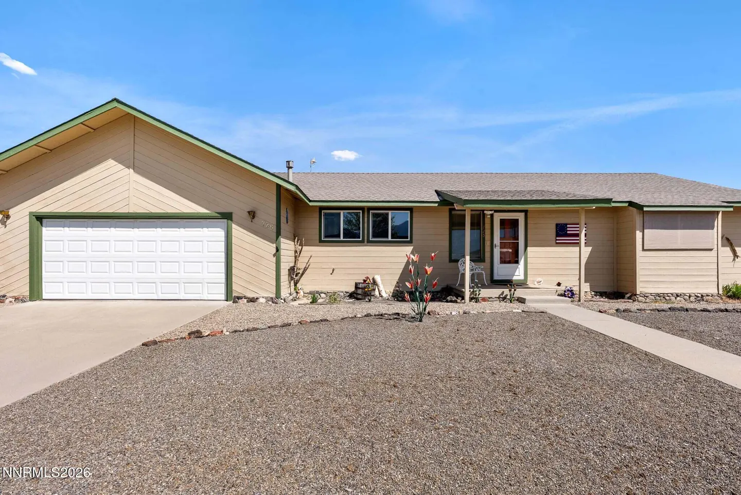 Tan single-story house with a white garage door, green trim, and a gravel yard under a blue sky.