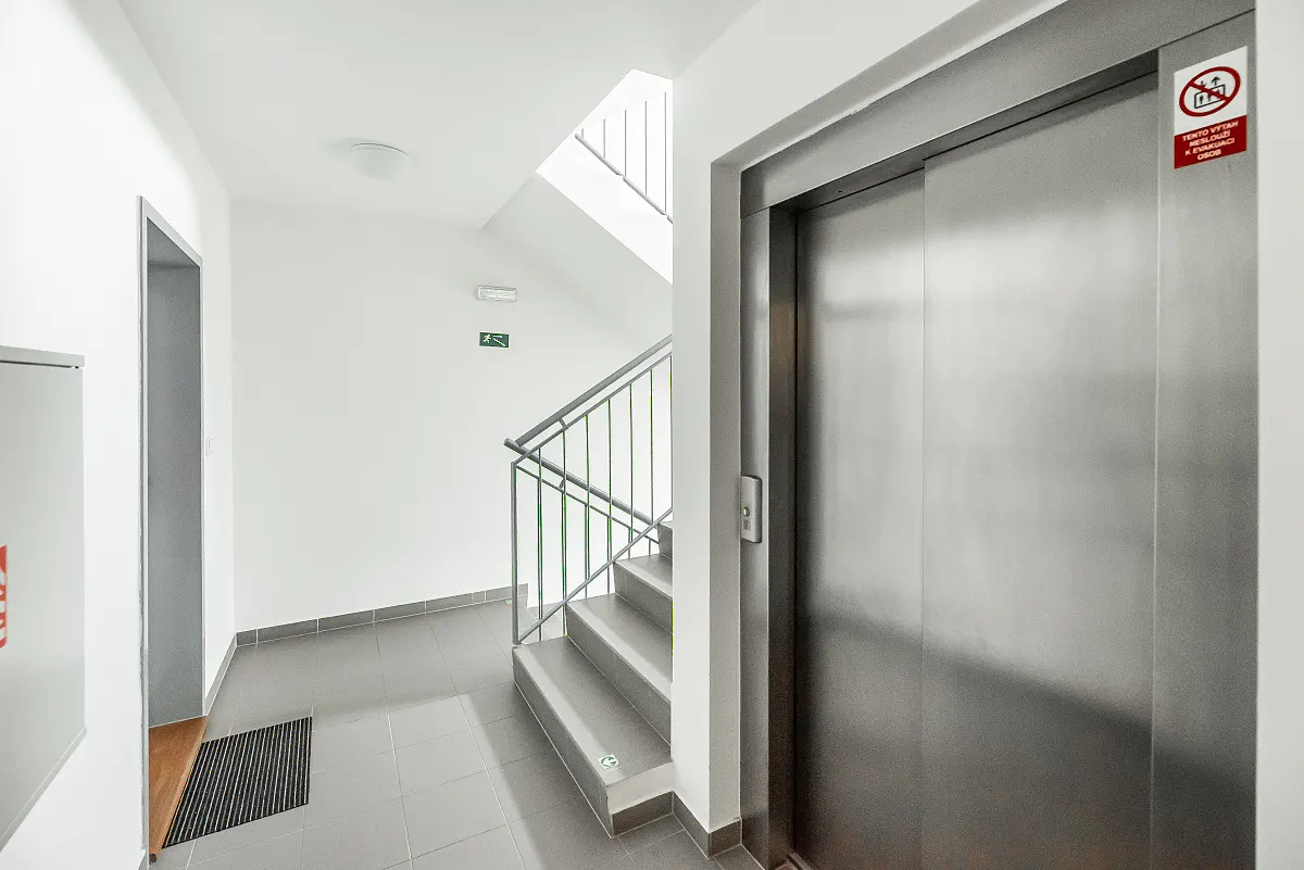 Modern apartment building hallway with a stainless steel elevator, grey tiled floor, and a staircase with metal railings.