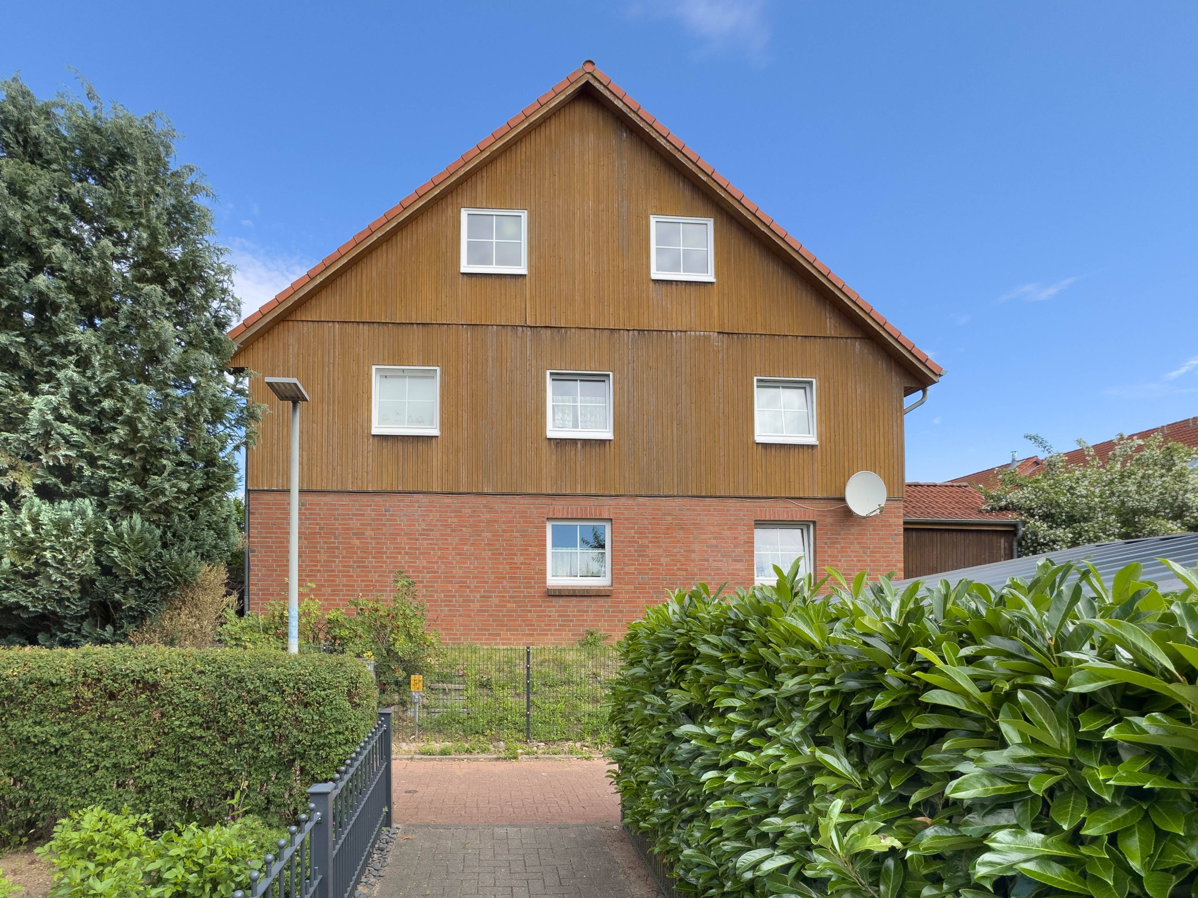 Two-story house with a brick ground floor, wood-paneled upper level, and gabled roof, framed by lush green hedges.