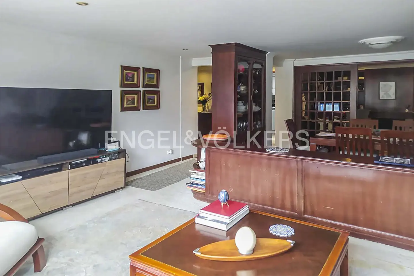 Living room with a large TV, wooden media console, coffee table with decor, and a dining area with a dark wood hutch.