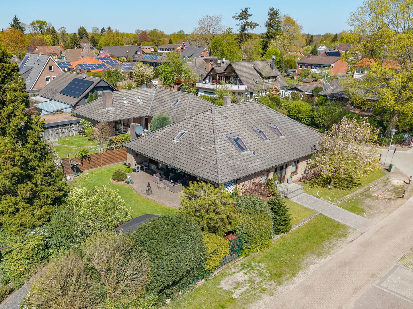 Aerial view of a brick house with a gray tiled roof, skylights, and a patio with outdoor furniture, surrounded by green trees and lawns.