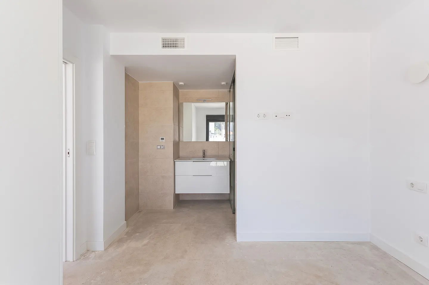 Modern white room with a floating vanity, mirror, and beige tiled wall, leading into an open-concept bathroom area.